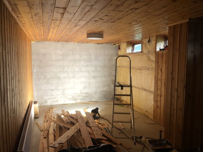 Renovering av källarrum med borttagen furupanel och invändig isolering, stege står i rummet och brädor ligger på golvet.