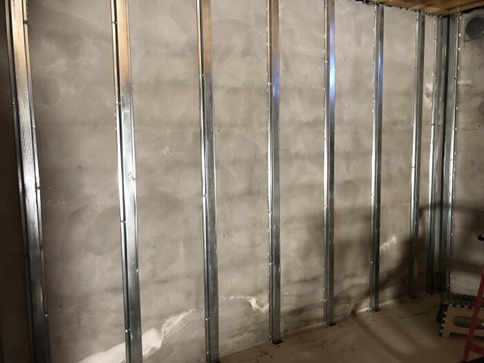 En vägg i en källare med flera vertikala metallreglar monterade på en betongvägg, förberedelse för installation av fibercementskivor, inga golv- eller takreglar.