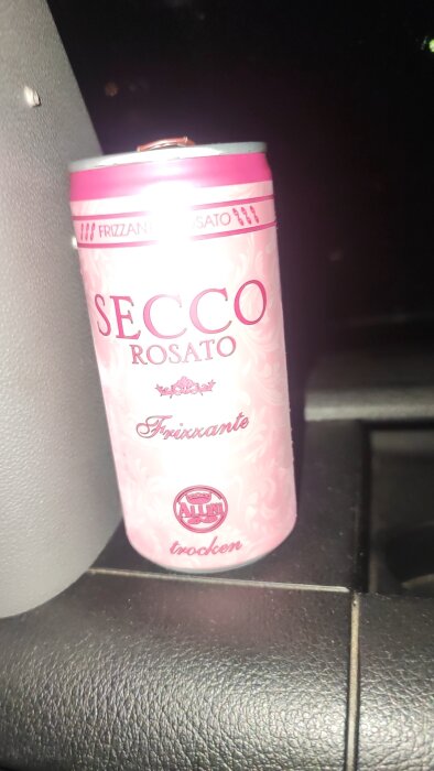 En rosa burk med märket "Secco Rosato Frizzante" placerad på en mörk yta.