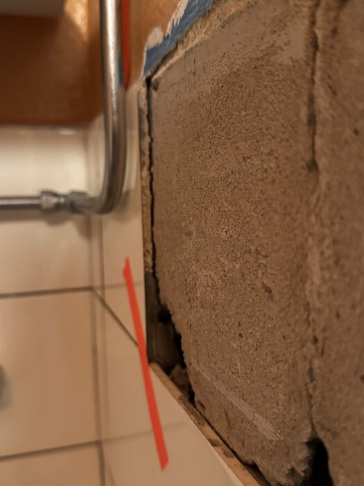En närbild av ett delvis avlägsnat lager av gammalt kakelfix på en vägg med synlig kant mellan kakel och det tjocka lagret fix. En metallrör är monterad på väggen.