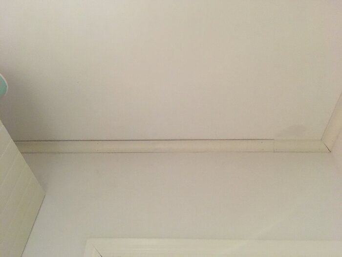 Glipa mellan vita taklister och tak i ett rum, där en liten del av en vägg också syns längst ner i bilden.