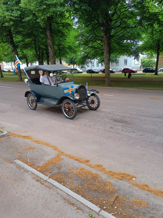 Vintage T-Ford med Ålandsflaggor kör längs en väg kantad av träd. Två personer i bilen vinkar.