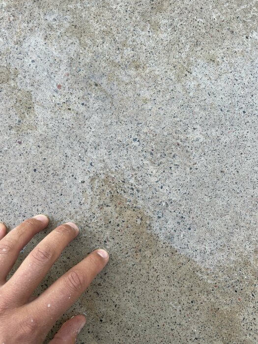 En hand som pekar på en betongyta efter behandling med högtryckstvätt. Ytan har en ojämn färg och struktur, vilket kan indikera kvarvarande betonghud.