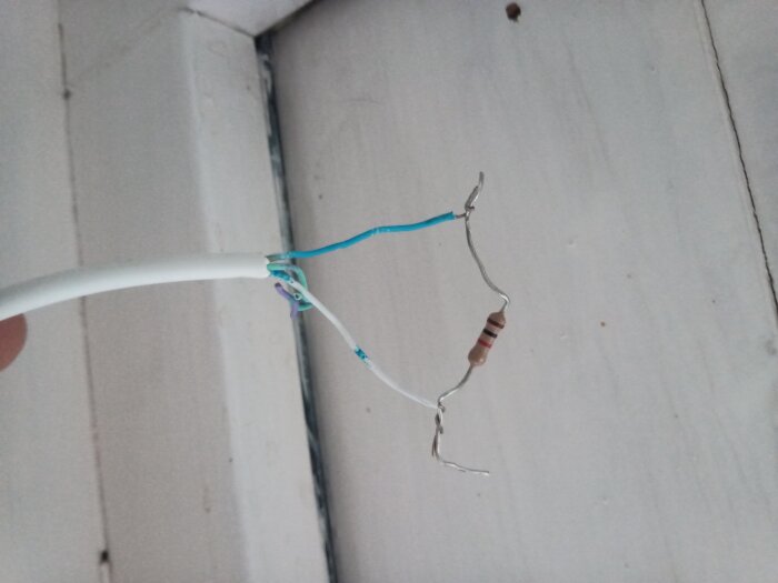 En vit kabel med två blå trådar som är kopplade till ett motstånd, hålls upp framför en vit vägg.