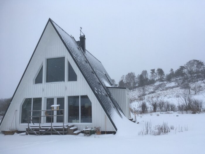 A-frame hus under byggnation i ett snöigt landskap, utsikt över en veranda och ett stort glasfönster i förgrunden.