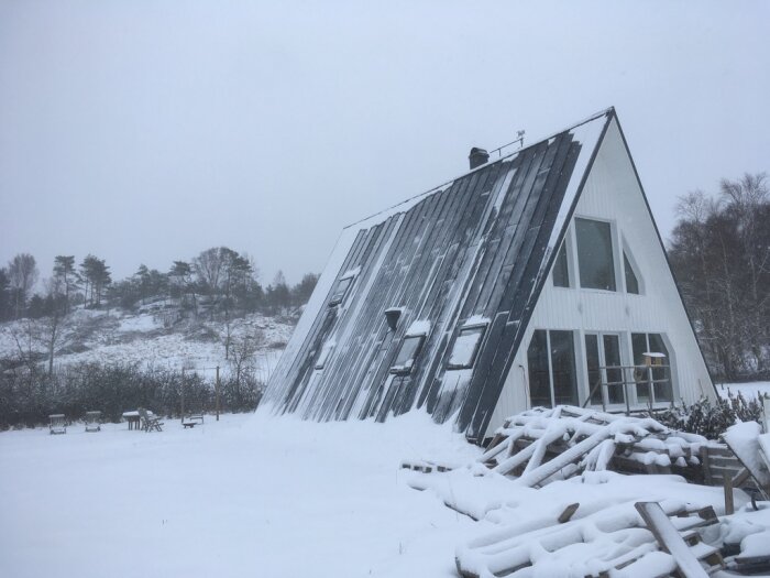 A-formad stuga med stora fönster täckt av snö i ett vinterlandskap, några byggnadsmaterial ligger framför huset.