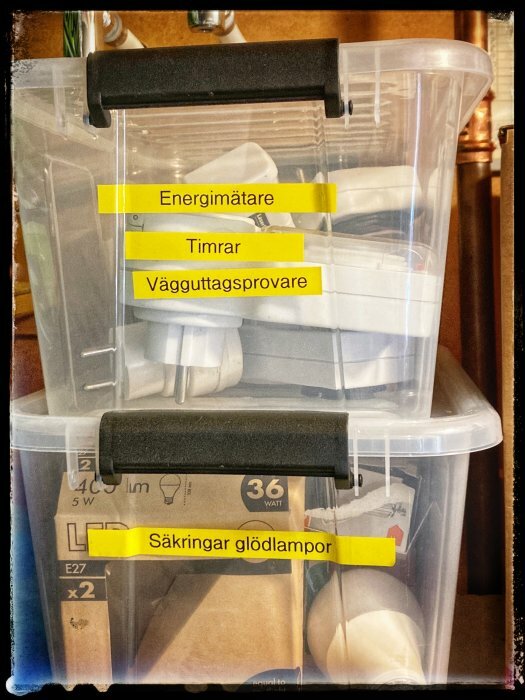 Två genomskinliga plastlådor med innehåll tydligt märkta med gula etiketter: "Energimätare", "Timrar", "VägguttagSprovare" och "Säkringar glödlampor".