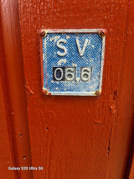 Närbild på en blå skylt med bokstäverna "SV" och siffrorna "06,6" på en röd trävägg.