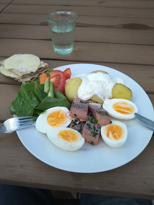 Tallrik med kokta ägg, potatis, gräddfil, gravad fisk, sallad och tomat på träbord, bredvid ett glas vatten och knäckebröd med ost.