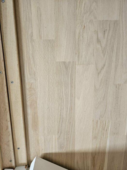 Nylackat trägolv visar ojämnheter i färg och droppar av lack, speciellt mot väggen. Ljusa träplankor med en vit sockel syns också.