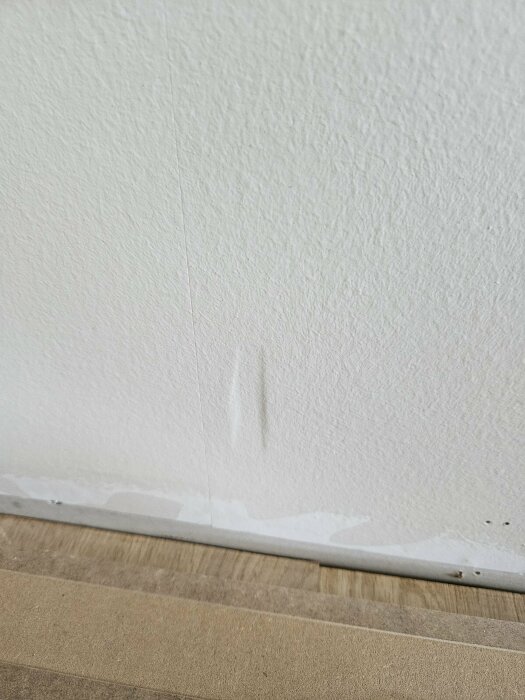 Skadad nymålad vit vägg vid golvlisten med märken av en slipmaskin och ojämn färg längs kanten. Golvet har nyligen blivit slipat och lackerat.
