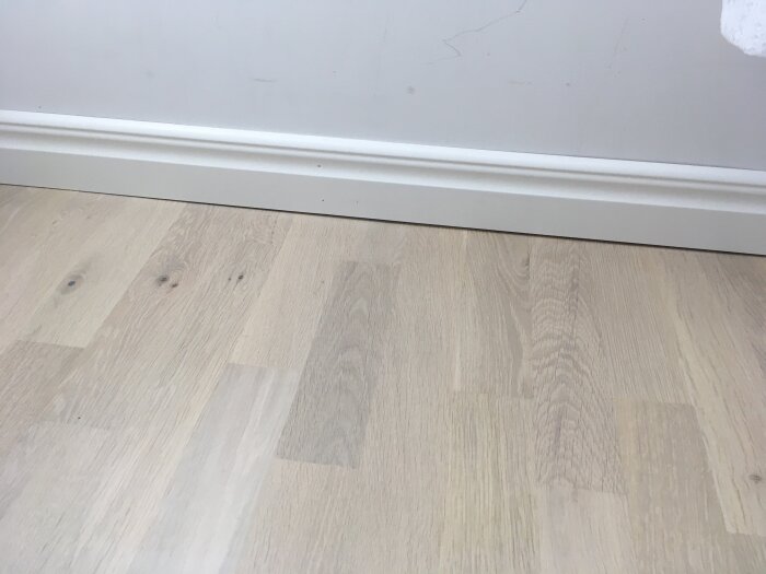Närbild av en golvlist som sitter ojämnt mot en vägg, med en glipa mellan listen och väggen. Golvet är av ljust trä med synliga ådringar.