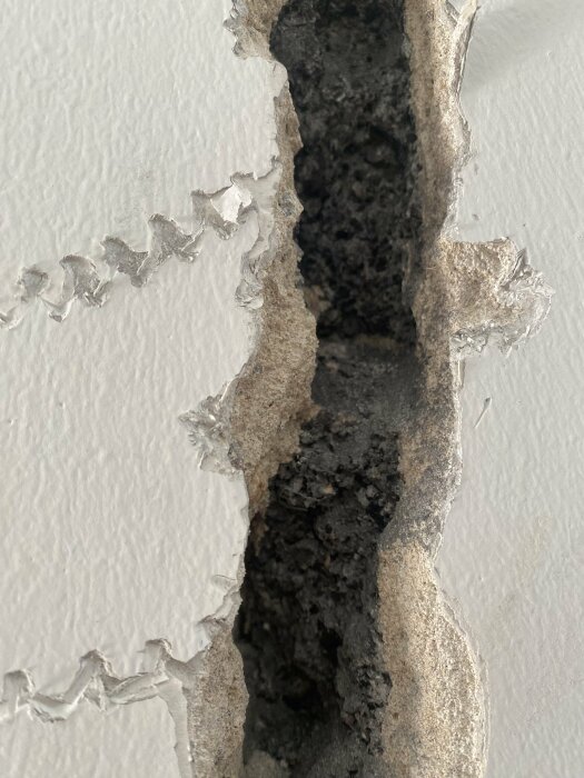 Bild på en murad innervägg med en öppning som avslöjar en mörk, smulig substans inuti, potentiellt misstänkt för att innehålla asbest.