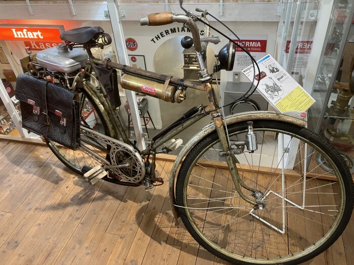 Gammal cykelutställning med svart läderväskor, stående på ett trägolv, omgiven av skyltar och föremål relaterade till teknikhistoria i ett museum.