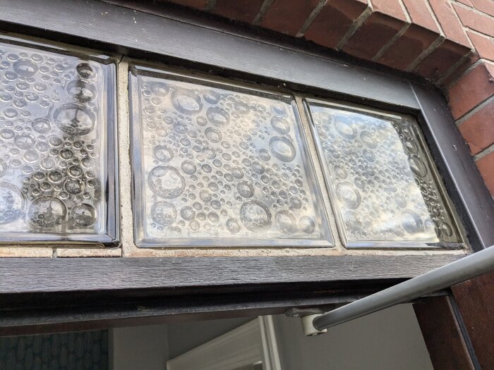 Fyra glasblock med bubbelmönster över en ytterdörr som behöver fogas om, belägna i en tegelvägg.