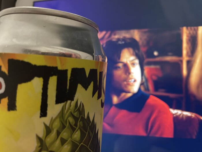 En ölburk med etiketten "Hoptimistic" framför en tv-skärm som visar en scen från filmen Bohemian Rhapsody.