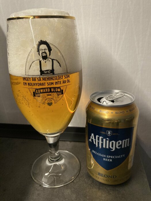 Ett glas av en belgisk öl med mycket skum bredvid en burk Affligem Blond belgiskt specialöl. Glaset har texten "Inget är så meningslöst som en kolhydrat som inte är öl.