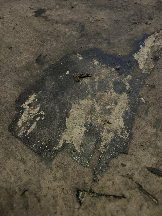 En detaljbild av en smutsig cementgolvyta med fläckar och märken, vissa partier ser oljiga eller fuktiga ut.