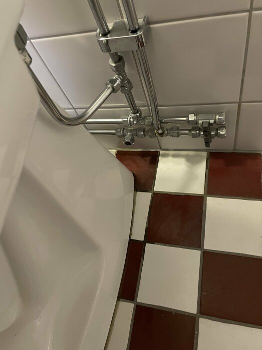 Toalett och rörsystem monterade mot vita kakelplattor på väggen, med fuktiga fogar på de rutiga kakelplattorna på golvet.