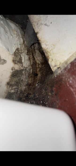 Bild av ett avlopp bakom en toalett med synligt vattenläckage och fuktfläckar på väggen och golv.områden, vilket tyder på problem med tätning.