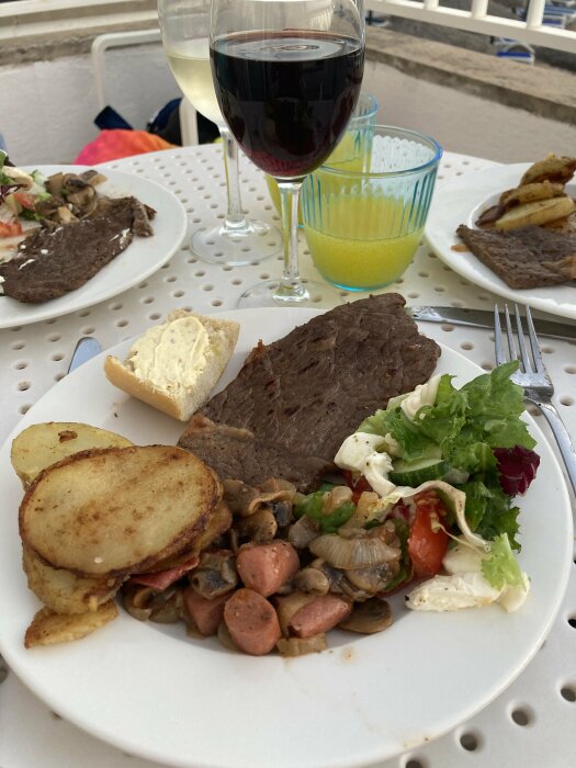 Tallrik med stekt kött, stekt potatis, svamp, korv, sallad och bröd. Två glas vin och ett glas juice på ett bord på en balkong.