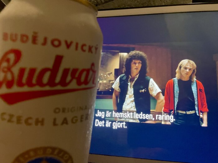 En burk Budweiser Budvar på ett bord, och i bakgrunden två män på en TV-skärm med texten "Jag är hemskt ledsen, raring. Det är gjort.