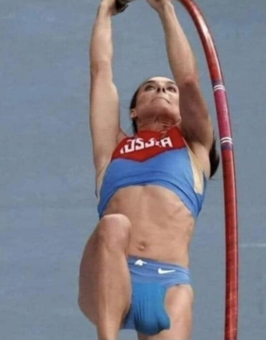 En stavhoppare i blåa och röda sportkläder hänger från en stav i luften, mitt i ett hopp.