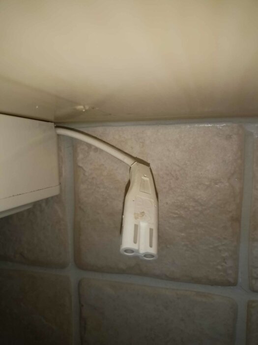 Vit tvåpolig kontakt hänger ut från en elkapsling monterad under ett köksskåp mot en tegelvägg.