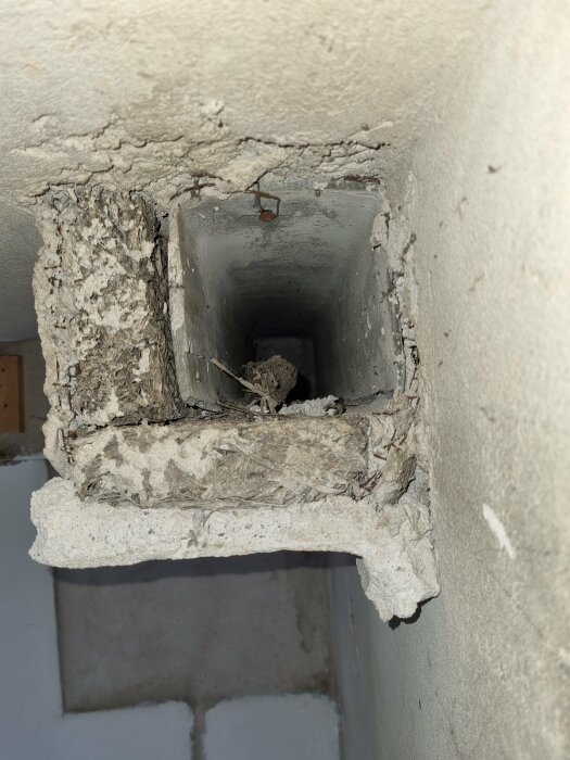 Ventilationskanal som sticker ut från väggen, delvis riven med synliga lager av cement, isoleringsmaterial och det inre röret, omgiven av sprucken puts.