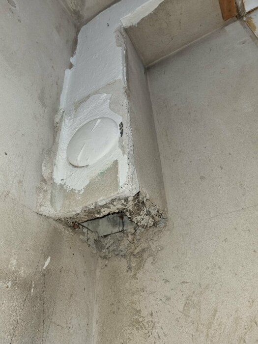Ventilationskanal i hörn av ett rum, delvis nerriven och täckt med cement. Synlig skada på kanalen, där inre rör och isolering exponeras.
