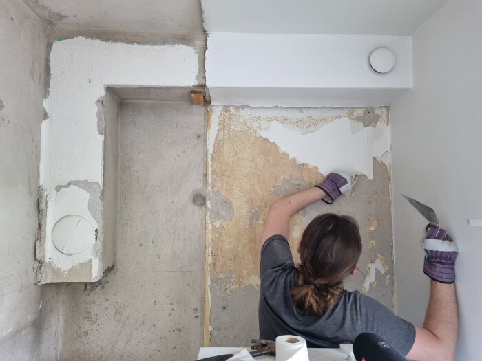 Person som renoverar ett kök från 50-talet, tar bort gammal tapet eller puts från en vägg nära en vitmålad L-formad ventilationskanal i hörnet av rummet.