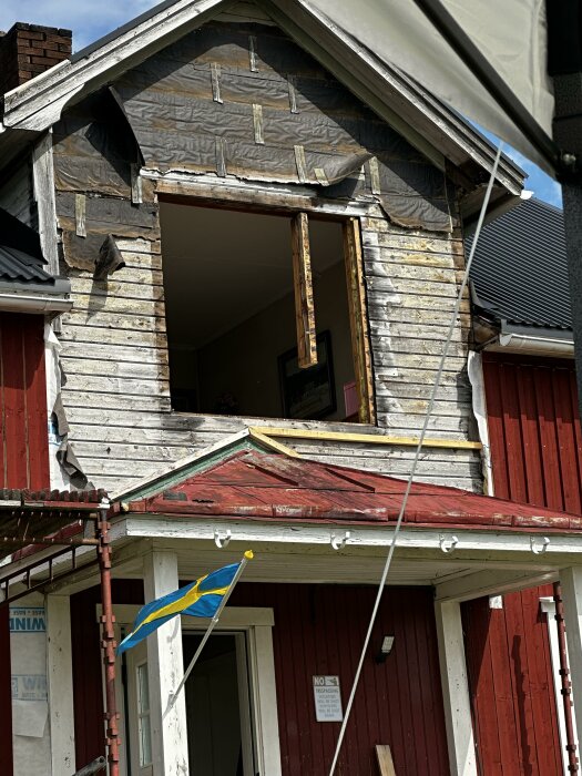 Ett rött hus med en övre våning där ett fönster har tagits bort, vilket avslöjar skadad stomme. En svensk flagga är monterad vid entrén.