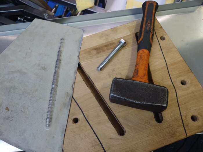 En handhållen hammare med svart och orange handtag, en bult och en metallbit med en svetsfog på ett arbetsbord.