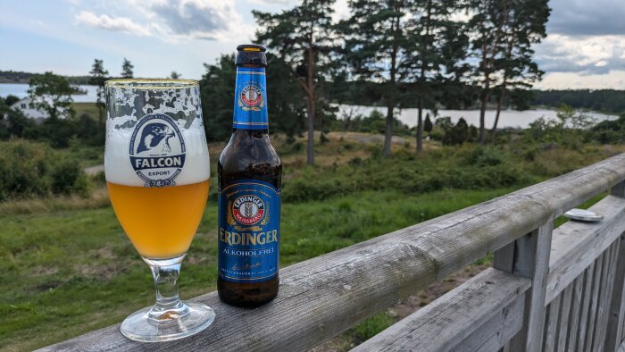 Ett glas Falcon öl och en flaska alkoholfri Erdinger på ett räcke med utsikt över en sjö och skog.