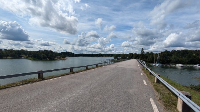 En bro över en sjö med båtar på höger sida och skogsområden på båda sidor under en himmel med moln.