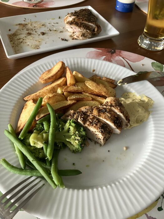 Tallrik med saftig kryddad kycklingfilé, klyftpotatis, haricots verts och broccoli, serverat med sås. ölglas syns i bakgrunden.