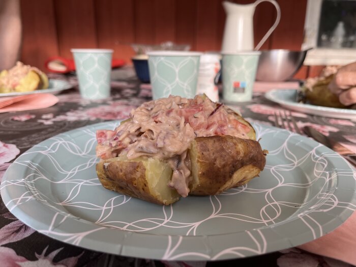 Bakad potatis med kycklingröra serverad på en mönstrad papperstallrik, med gröna engångsmuggar i bakgrunden och ett matsalsbord dukat.