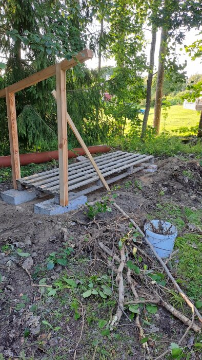 Vedförråd under uppbyggnad med en träställning på cementblock. Ogräs har rensats och en blå hink med ogräsrötter står framför ställningen.