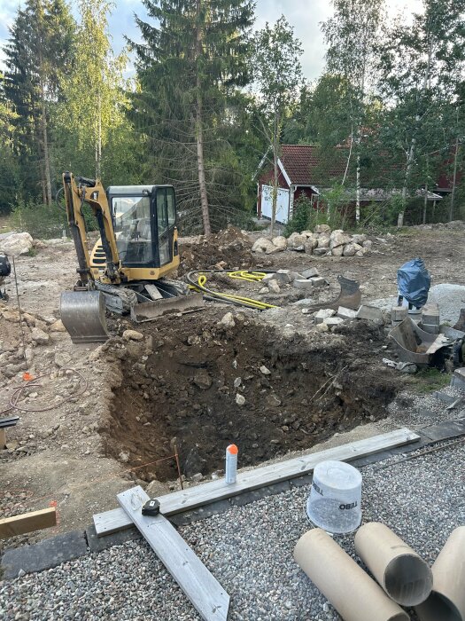 En gul grävmaskin står i ett schaktat område omgiven av jord och stenar, med en skog och en röd stuga i bakgrunden.