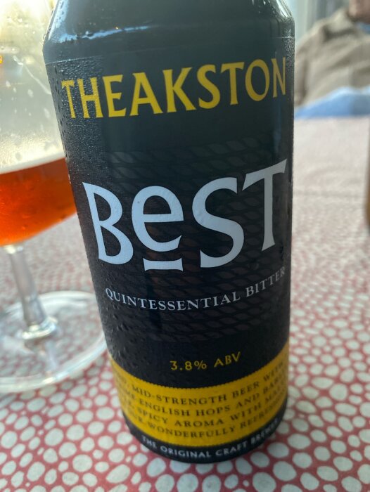 En burk Theakston BEST Quintessential Bitter öl med 3.8% alkoholstyrka står på ett bord bredvid ett glas öl.