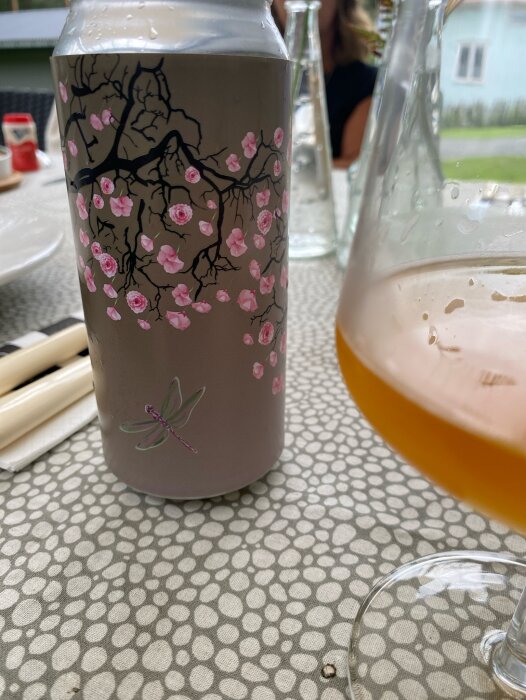 Ölburk med blommönster och en färgglad slända samt ett glas öl, placerade på ett bord utomhus under en ölprovning med vänner.