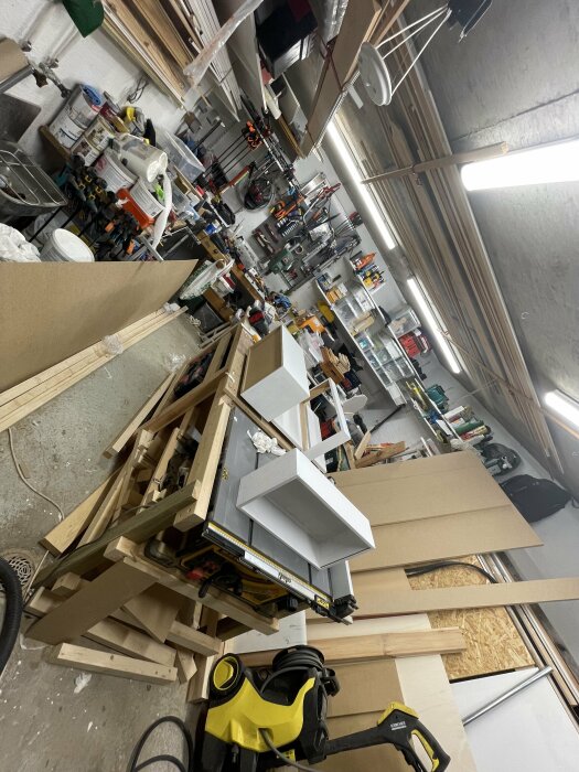 Två vitmålade kökslådor står på ett arbetsbord i en verkstad fylld med verktyg och byggmaterial.