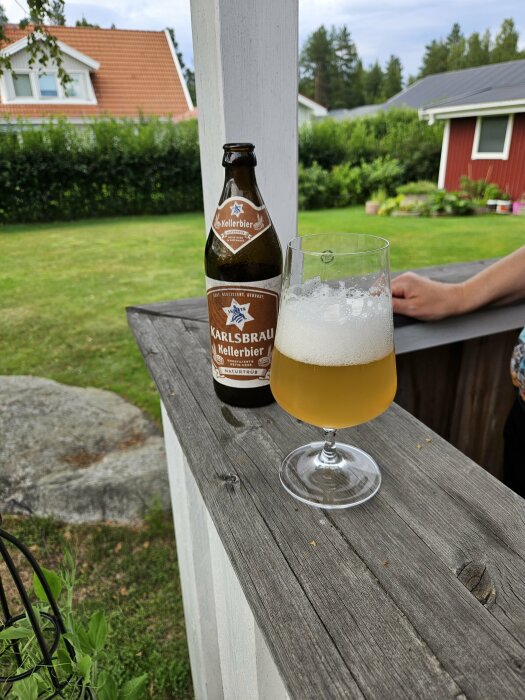 En ölflaska med etiketten "Karlsbrau Kellerbier" och ett fyllt ölglas står på ett trästaket i en trädgård med hus i bakgrunden.