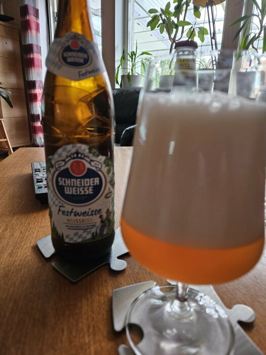 En flaska Schneider Weisse Festweisse Weissbier och ett glas med öl, stående på ett bord med underlägg i ett vardagsrum.
