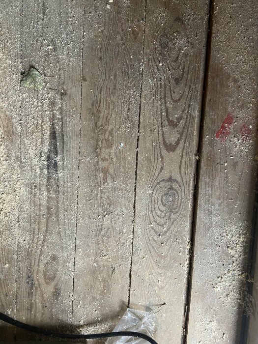 Träplankor på ett golv i ett förråd med tecken på smuts och eventuellt mögel, omgivna av skräp och sågspån.