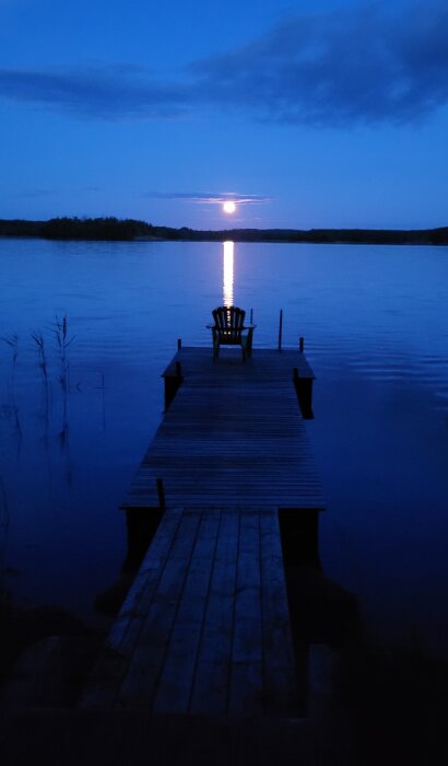 En brygga leder ut mot en sjö under nattid, med en stol i slutet och en stor måne som reflekteras på vattnet.