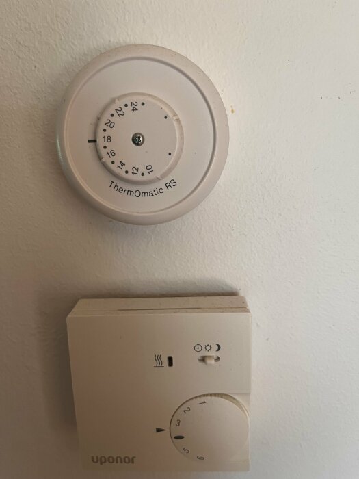 Två gamla termostater på en vit vägg. Den övre är rund och märkt ThermOmatic RS. Den undre är fyrkantig och märkt Uponor med en vredskiva.