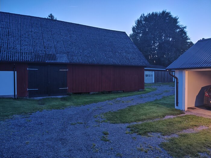 Röd ladugård med stor svart port till vänster och garage på höger sida med en bil parkerad, grusväg och gräs i förgrunden, träd i bakgrunden.