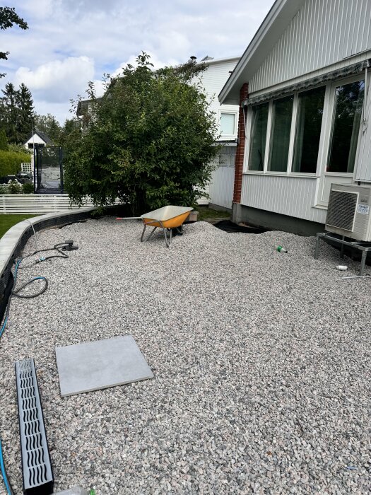 Uteplats med lager av makadam 8-16 mm, en skottkärra och en platta av granitkeramik 60x60 cm ligger på marken, omgivet av husväggar och träd.