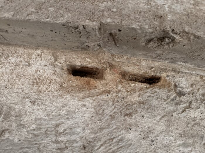 Två närliggande hål i betonggolvet i en gillestuga, med tecken på gammalt trä och fuktskador.